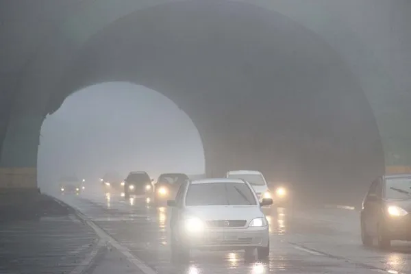 Carros com faróis acesos saindo de um túnel numa rodovia coberta de neblina